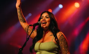 A tattooed woman sings into a microphone on a dimly lit stage, her arm raised.