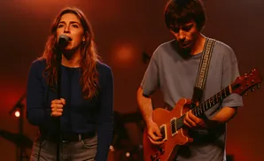 A woman sings into a microphone while a man plays guitar onstage.