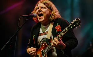 A male guitarist sings into a microphone with his eyes closed and blonde hair flowing.