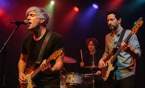 A band performs on stage with a singer and guitarist in the foreground, and a bassist and drummer in the background.