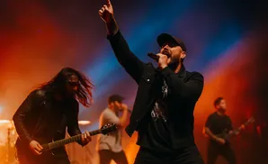 A lead singer points to the sky with one hand while singing into a microphone, performing with his band.