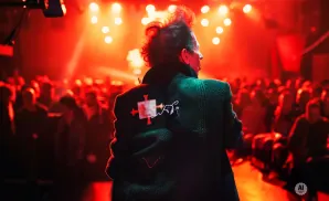 Man with spiky hair on stage with red lighting and a crowd.