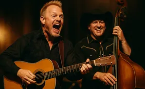 Two musicians perform on stage. A singer strums an acoustic guitar while a man in a cowboy hat plays the upright bass.