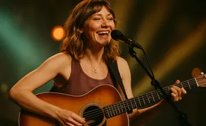 A woman with a guitar laughs and sings into a microphone on stage.