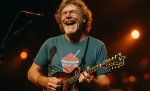 A laughing man with glasses plays a mandolin on stage.