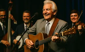 Man in a gray suit happily plays an acoustic guitar, with other musicians in the background.