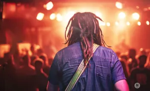 Man with dreadlocks and a denim shirt stands with his back to the camera at a concert.