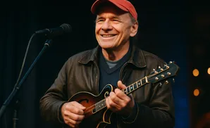 Man wearing a red cap and brown leather jacket smiles while playing a mandolin near a microphone.