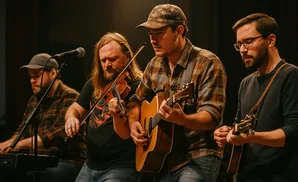 Four men playing musical instruments on a stage: a keyboardist, a violinist, a guitarist, and a mandolin player.