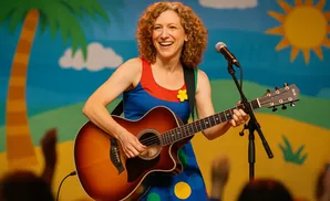 A woman with curly hair smiles and plays an acoustic guitar on stage, wearing a colorful dress.