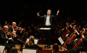 Conductor leading an orchestra with cellos and violins in front of a dark background.