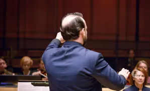 A conductor with thinning hair stands with his back to the camera, conducting an orchestra.