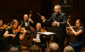 Conductor leads an orchestra with a violinist and flutist in the foreground.