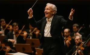 An elderly male orchestra conductor in a tuxedo smiles joyfully while leading musicians.