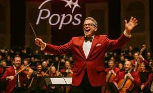 A conductor in a red jacket laughs with his arms outstretched, directing an orchestra.