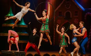 Circus performers in festive costumes perform aerial acrobatics and contortion with hula hoops and a backdrop of a gingerbread house.