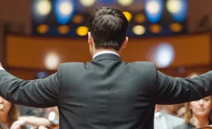 A conductor in a black suit faces away from the camera, arms outstretched, leading an orchestra.