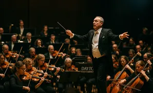 A conductor directs an orchestra with a baton, leading musicians playing violins and cellos.