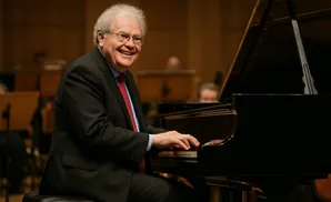 A smiling man in a suit plays a grand piano during a concert.
