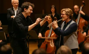 A conductor leads a female flute player in an orchestra, with a cellist and bassist in the background.