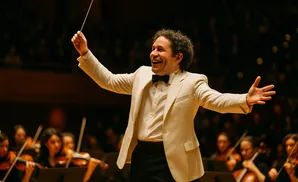A conductor in a cream suit leads an orchestra with a baton, smiling broadly with arms outstretched.