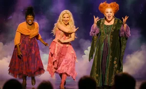 Three women in witch costumes dance on stage with purple lighting and fog effects.