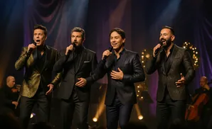 Four men in suits sing into microphones on a stage with purple curtains and warm lighting.