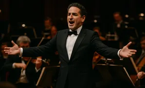 A male opera singer in a tuxedo, with arms outstretched, sings into a microphone with an orchestra behind him.