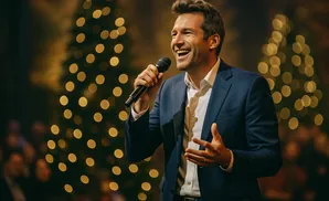 A man in a blue suit sings into a microphone, smiling, with blurred Christmas trees and lights in the background.