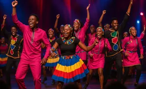 A choir in bright African-inspired attire sings and dances on stage with their arms raised.