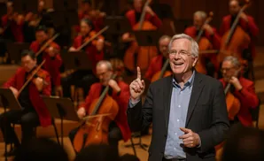 A conductor smiles while pointing up, with an orchestra in red jackets playing cellos and violins behind him.
