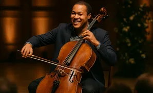 A Black man smiles while playing the cello in a concert hall.