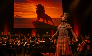 A performer in a regal costume stands on stage before a screen displaying a lion and an orchestra.