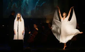 A young woman in a white hooded robe sings into a microphone, while a dancer in a white dress performs to the side.