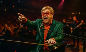 Elton John, wearing a sparkly green jacket and orange shirt, plays piano and points at the audience.