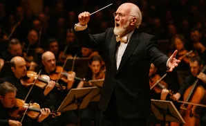 Conductor John Williams leads an orchestra, baton raised in his right hand, conducting with passion.