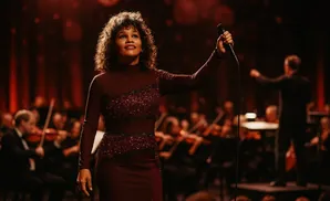 Singer with curly hair in a sequined dress holding a microphone on stage with an orchestra.