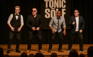 Four men in formal wear stand on a stage, performing.