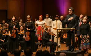 A conductor and orchestra members stand on stage in formal wear and traditional costumes.