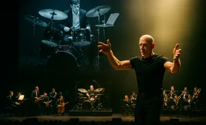 A conductor leads a jazz band while a drummer plays on a large screen behind them.