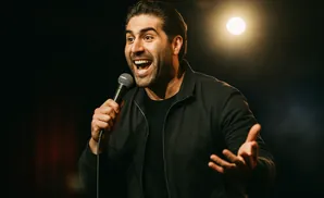 A man with dark hair holds a microphone and speaks into it while gesturing with his hand.