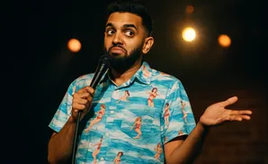 A man with a beard holds a microphone and gestures with his hand, wearing a Hawaiian shirt with a beach scene pattern.