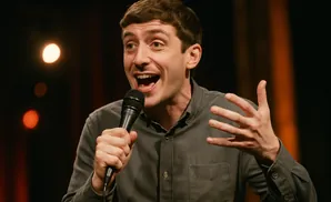 A man with brown hair speaks into a microphone with an open mouth, gesturing with his hand.
