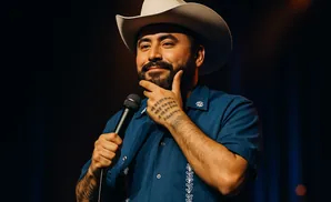A man in a cowboy hat and blue shirt holds a microphone and rests his chin on his hand.