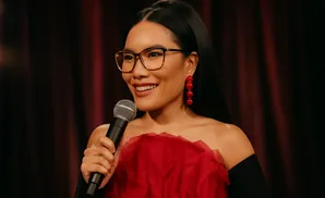 A woman with glasses and red earrings smiles while holding a microphone, wearing a red top and black sleeves.