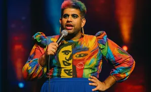A person with rainbow hair and vibrant clothing holds a microphone and stands on stage.