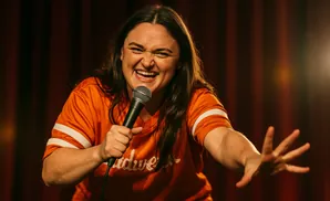 A woman in an orange t-shirt stands with a microphone, laughing and gesturing with her hand toward the right.