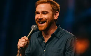 A ginger-haired man with a beard smiles broadly, holding a microphone and wearing a blue collared shirt.