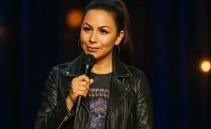 A woman with dark hair in a ponytail, wearing a black leather jacket, holds a microphone.