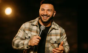 A smiling man in a plaid jacket holds a microphone and gestures with his other hand.